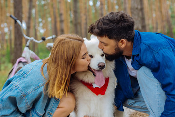 Kissing dog. Cute young couple wearing denim clothes kissing their white fluffy dog walking in the park