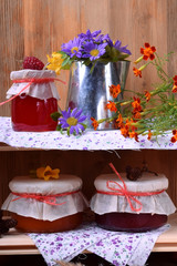 Jars of different jam and a metallic jug with flowers on wooden shelves