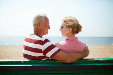 Aged man embracing his wife while both sitting on bench by seaside and looking at each other