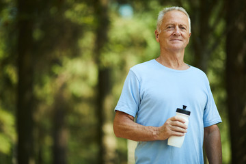 Sporty senior man with bottle of water standing in park or forest on summer day and having...