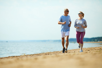 Active senior man and woman running down sandy beach with waterside on background