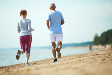 Back view of senior couple in activewear running along coastline on sandy beach