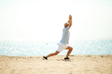 Active retired man in sportswear stretching legs and keeping his raised hands together while exercising on the beach