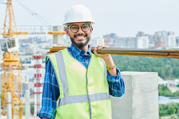 Waist up portrait of cheerful Middle-Eastern construction worker wearing harhat and holding metal...