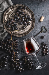 Red wine concept with glass, grapes in old plate and vintage corkscrew on dark table background,...
