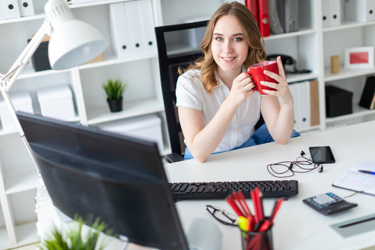Beautiful young girl working with computer in office. The girl has a red cup in her hands.
