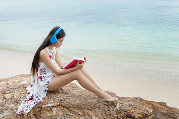 Beautiful smiling girl listening to music and reading a book on the beach of blue ocean and sky on the background.