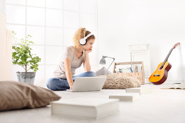 young woman with computer, headphones, guitar and books, sitting on the floor in living room on white wide window in the background