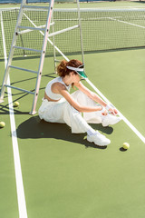 side view of young woman in stylish white clothing and cap tying shoelaces on tennis court with racket and balls