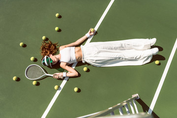 overhead view of stylish woman in white clothing and cap lying with racket lying on tennis court with racket
