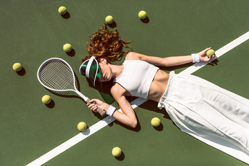 overhead view of stylish woman in white clothing and cap lying with racket lying on tennis court...