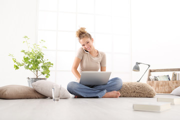 smiling young woman with computer talk on the cell phone, sitting on the floor in living room on white wide window in the background
