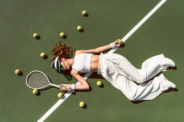 overhead view of attractive woman in white clothing and cap lying with racket lying on tennis court with racket