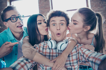 Close up portrait of cheerful youngsters make a selfie on front of the smartphone girls in casual wear kiss the guy on the cheek plump and pouted lips