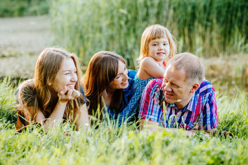 Fototapeta premium Ideal picture of family happiness - happy family with two daughter having fun and lying on the grass in lake shore on a summer day in sunset. Parenthood, love, vacation and happy family concept.