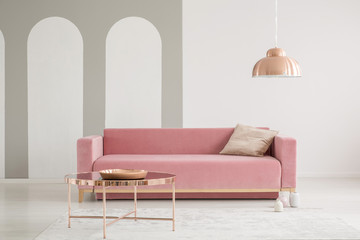 Copper table in front of pink sofa with cushion in white living room interior with lamp. Real photo