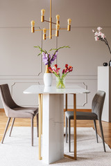 Flowers on table in grey dining room interior with gold lamp and chairs on carpet. Real photo