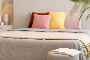 Book on stool next to grey bed with blanket and colorful pillows in bedroom interior. Real photo with focus on the blanket