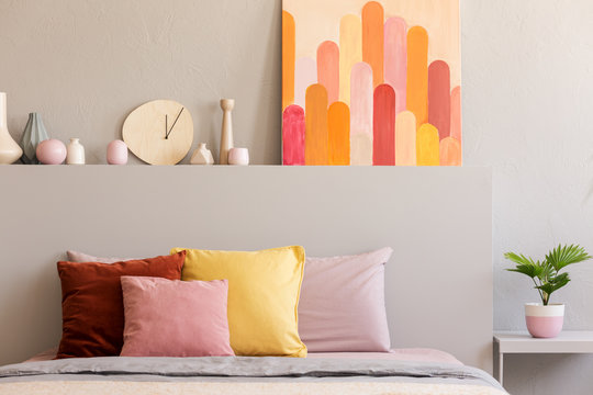 Colorful pillows on bed in grey bedroom interior with poster and clock on bedhead. Real photo