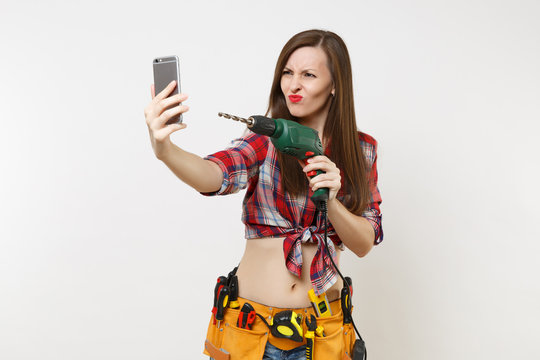 Strong excited handyman woman in shirt, kit tools belt full of instruments doing selfie on mobile phone with power electric drill isolated on white background. Female in male work. Renovation concept.