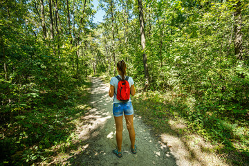 Naklejka na ściany i meble Young girl backpacker in jeans shorts with red backpack and sandals on his feet, walks on the road in the green forest in the sunshine outdoors in the summer time