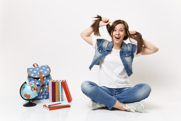 Portrait of young crazy amazed woman student in denim clothes holding ponytails, sitting near globe, backpack, school books isolated on white background. Education in high school university college.