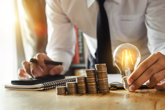 business man hand holding lightbulb with using smartphone and calculator to calculate and money stack. idea saving energy and accounting finance in morning light 