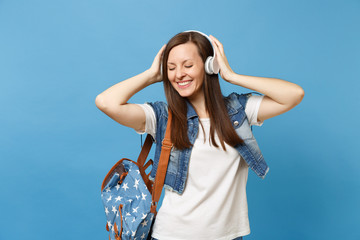 Young joyful woman student with closed eyes in denim clothes with backpack keeping hand on headphones listen music isolated on blue background. Education in university. Copy space for advertisement.