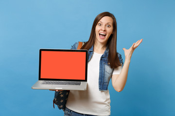Young excited woman student with opened mouth spreading hands holding laptop pc computer with blank black empty screen isolated on blue background. Education in college. Copy space for advertisement.