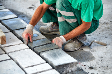 A workman's gloved hands use a hammer to place stone pavers. Worker creating pavement using cobblestone blocks and granite stones.