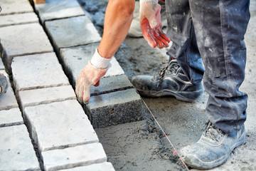 A workman's gloved hands use a hammer to place stone pavers. Worker creating pavement using cobblestone blocks and granite stones.