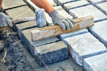 A workman's gloved hands use a hammer to place stone pavers. Worker creating pavement using cobblestone blocks and granite stones.