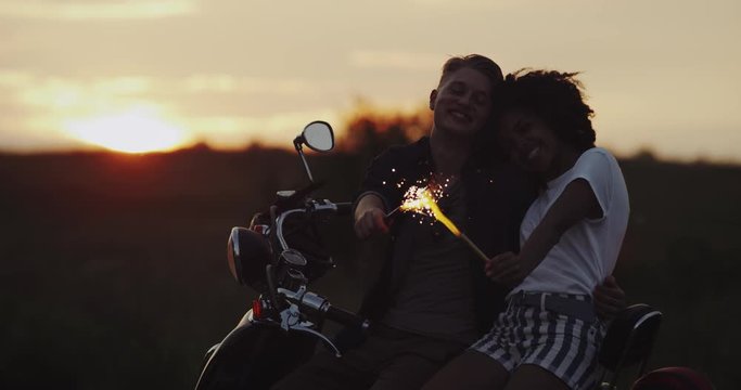Beautiful young woman and man stand on their retro motorbike holding the sparklers and enjoying the time at sunset.