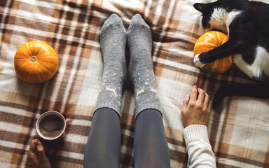 Fototapeta na wymiar Women's hands and feet in sweater and woolen cozy gray socks holding cup of hot coffee, sitting on plaid with kitten, pumpkin, candles and leaves. Concept winter comfort, morning drinking.