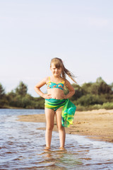 little joyful blonde with a tail girl in a swimsuit and a pareo posing and jumping on the river bank in sunny summer weather