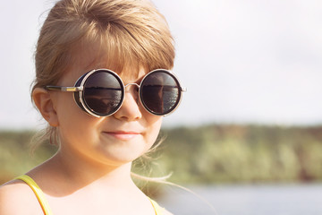 Fototapeta na wymiar Portrait of a small blond girl with glasses on the river bank in sunny summer weather