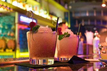 Two alcoholic cocktails of pink color with raspberries, ice and mint are on bar. Neon lighting. The concept of Friday's holiday.