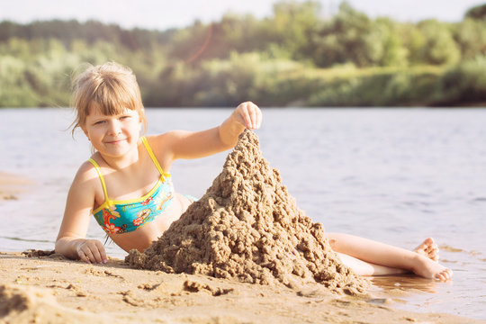 a small blonde joyful girl in a swimsuit is building a sand castle on the river bank in sunny warm weather in the summer