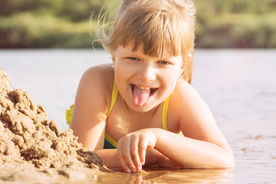 a small blonde joyful girl in a swimsuit builds a sand castle on the river bank in sunny warm weather in the summer. portrait