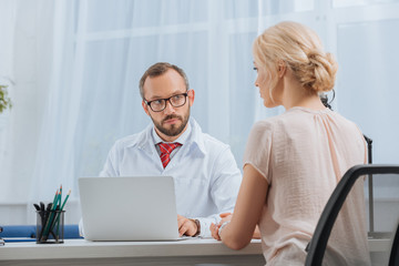 physiotherapist in white coat having conversation with female patient at workplace with laptop in...