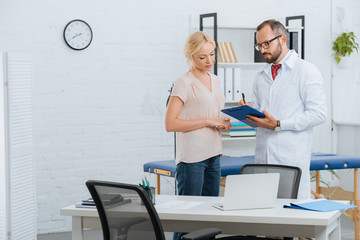 Obraz premium portrait of chiropractic in white coat with notepad and female patient having conversation in clinic