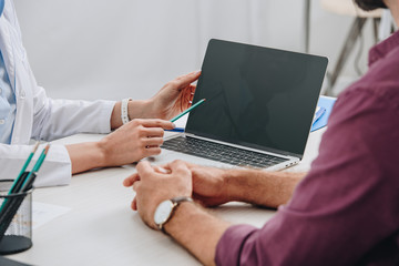 partial view of doctor pointing at laptop screen with patient near by at workplace in hospital