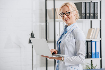 side view of smiling chiropractor in eyeglasses and white coat with laptop in clinic