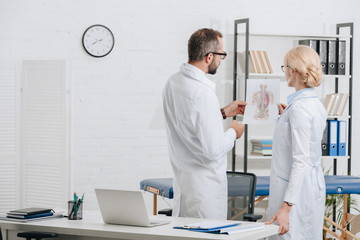 Obraz premium back view of physiotherapists in white coats and eyeglasses discussing work in clinic