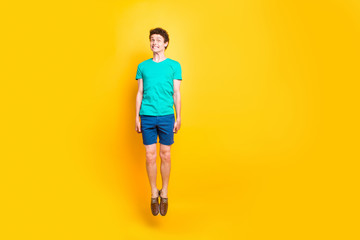 Fototapeta na wymiar Full size length body picture of handsome curly-haired playful young guy wearing casual green t-shirt, shorts, shoes, jumping up in air, grimacing. Isolated over yellow background