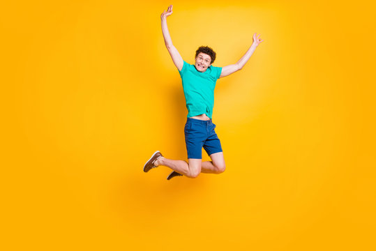 Young attractive handsome funky curly-haired excited guy wearing casual green polo t-shirt, blue shorts, sneakers, flying in air, raising hands up. Isolated over vivid shine bright yellow background
