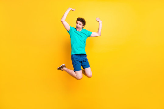 Full size length body picture of handsome curly-haired playful young guy wearing casual green t-shirt, shorts, shoes, jumping in air, hands up, party time. Isolated over yellow background