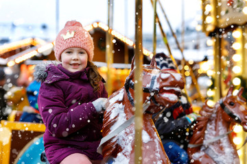 Adorable little kid girl riding on a merry go round carousel horse at Christmas funfair or market,...