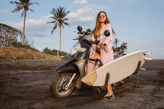attractive smiling girl sitting on scooter with surfboard on beach in bali, indonesia