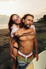 Fototapeta premium smiling girlfriend hugging boyfriend with surfing board on beach in bali, indonesia